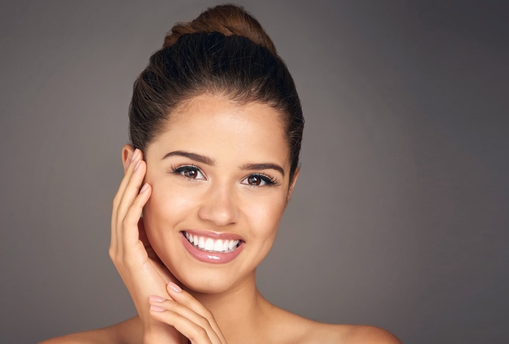 Woman in studio with glowing skin smiling and touching face, concept of clean skincare, natural beauty, and dermatology self-care on gray background.